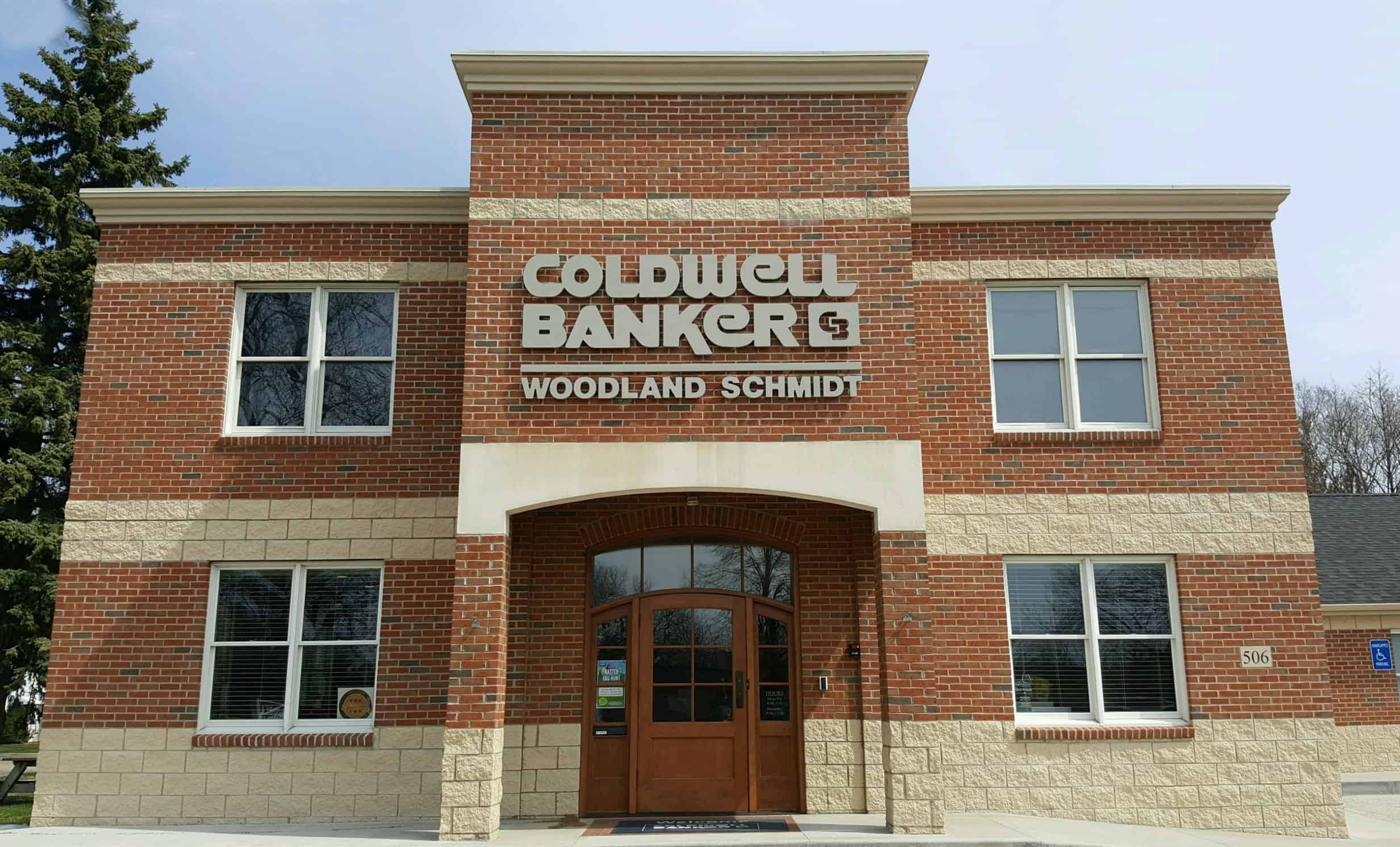 Modern commercial building with brick exterior, featuring Coldwell Banker real estate signage and Woodland Schmidt branding, serving as a local real estate office.