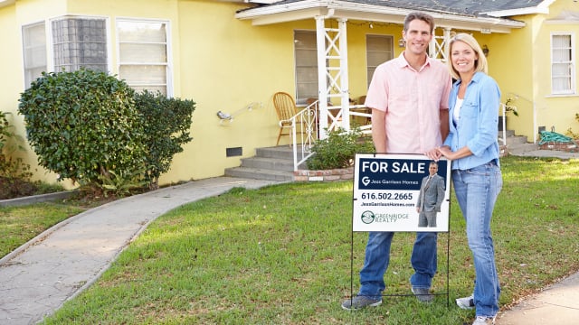 Beautiful couple standing in front of a charming yellow home with a for sale sign, showcasing Jess Garrison Homes' real estate expertise and successful home sales.
